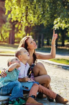 Young mother sitting with her children in the park, showing them something above with finger, they are looking with interest.