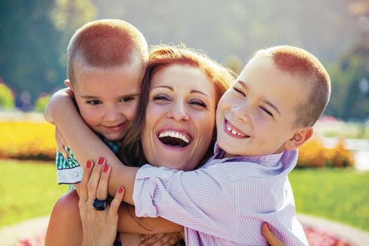 Young mother being embraced by her two little sons laugh infront of colorful summer flowers in a park.