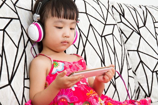 Chinese little girl on headphones holding mobile phone sitting on sofa