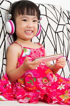 Chinese little girl on headphones holding mobile phone sitting on sofa