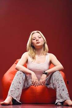 A cute girl in pajamas sitting on a red beanbag chair isolated on red background