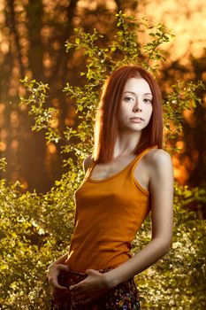 Beautiful redhead girl casually walking under the rays of the sunset in summer forest