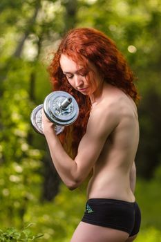 fitness, health, diet concept - stunning beautiful redhead girl with long curly hair and great musculature exercising with heavy dumbbells at summer spring forest