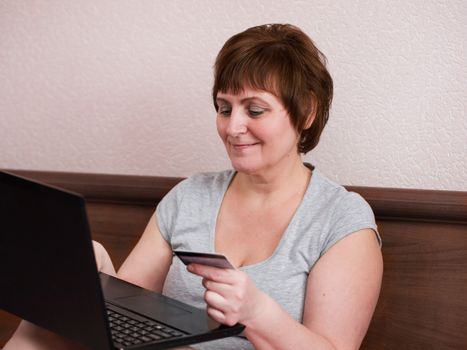 Smiling senior adult woman sitting at bedroom with laptop and holding bank card in her hand