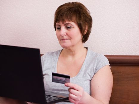 Portrait of senior woman sitting at bedroom with her laptop and  holding bank card in her hand, senior adult woman makes a purchase online.