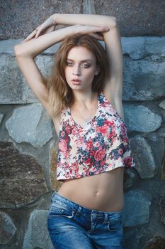 Young sexy woman model in jeans and tank top posing for fashion portrait near old weathered wall outside