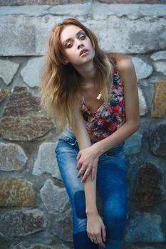 Young sexy woman model in jeans and tank top posing for fashion portrait near old weathered wall outside