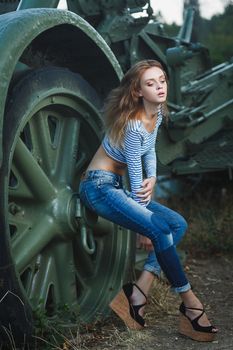 Young sexy woman model in jeans and striped tank top posing for fashion portrait near old artillery gun outside