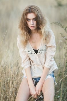 Young sexy woman model in jeans shorts and blouse posing for fashion portrait outside in a field at golden hour sunset