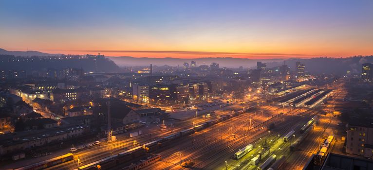 Cityscape of Slovenian capital Ljubljana at dusk. Aerial view of illuminated train tracks of a main railway station can be seen in forground.