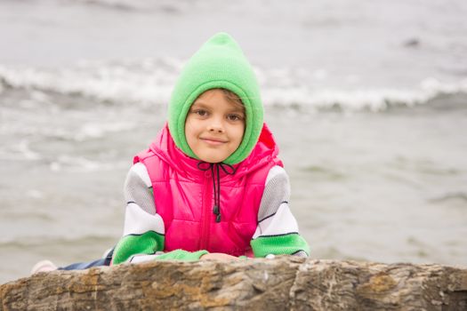 Warmly dressed girl looks out from behind a rock against the backdrop of the sea on a cold cloudy day
