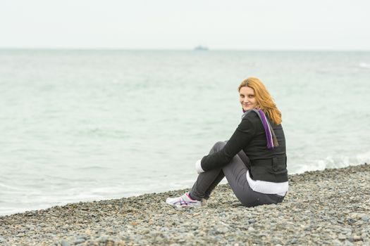  Smiling young girl sitting on a pebble beach by the sea on a cloudy cold day embracing knees