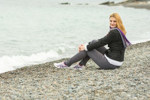 Joyful young girl sitting on a pebble beach by the sea on a cloudy cold day