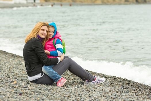 Mom and daughter in the arms sit on the beach on a cool day and watching the surf