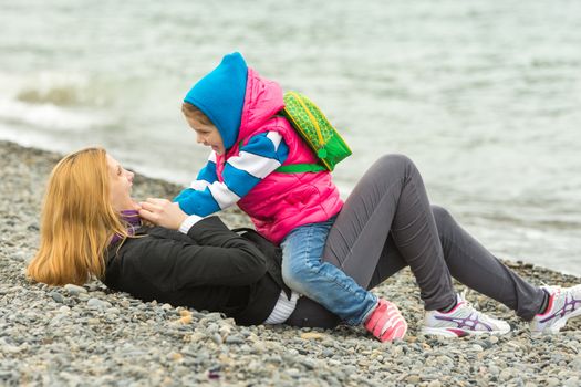 Five-year girl in warm clothes sits astride the mother who is lying on the pebble beach and sea fun looking at each other