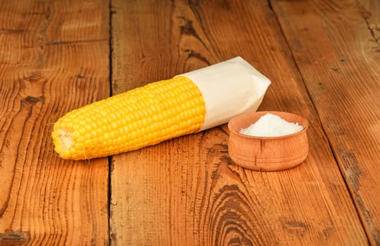 One open fresh yellow corn cob and salt in cup on brown vintage wooden surface