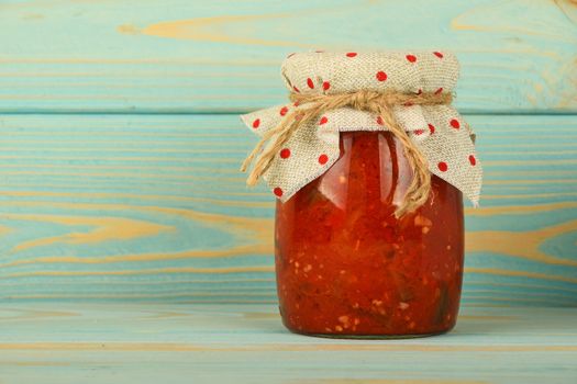 One glass jar of homemade pickled pepper, paprika and eggplant salad with dotted textile top decoration at blue painted vintage wooden surface