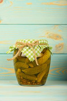 One glass jar of homemade pickled cucumbers with green checkered textile top decoration at blue painted vintage wooden surface