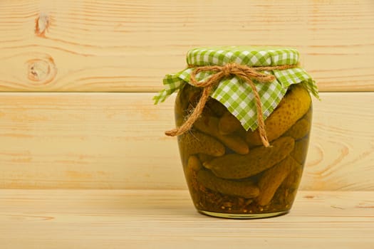 One glass jar of homemade pickled cucumbers with green checkered textile top decoration at beige painted vintage wooden surface