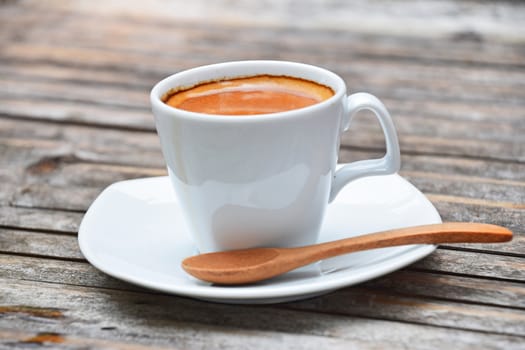 One white cup full of espresso coffee with brown crema on porcelain saucer with wooden spoon on old vintage bamboo table