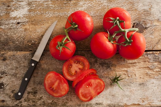 Preparing sliced ripe red tomatoes on a rustic wooden kitchen counter with a paring knife, overhead view of sliced and whole tomatoes on the vine