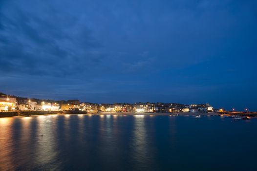 Scenic view of St Ives, Cornwall, UK at night with the lights reflected in the calm water of the bay under a twilight blue sky