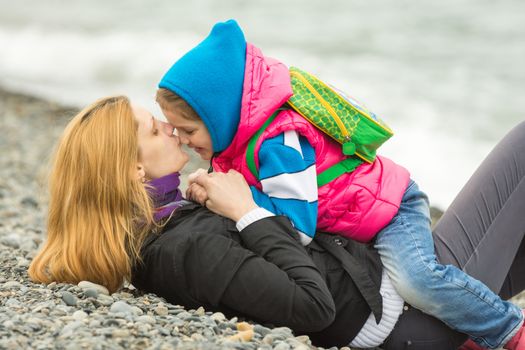 Young mother lying on the pebbly seashore kissing the nose her daughter sitting on her riding