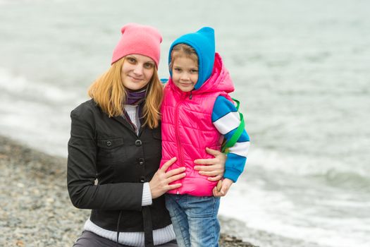 Mom and daughter in warm clothes hugging each other on the beach in cold weather and with a smile look in the frame