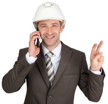 Smiling businessman in helmet talking on mobile and showing peace sign isolated on white background