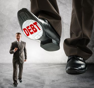 Big foot with word debt steps on businessman on grey wall background