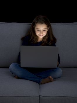 Happy little girl sitting on a couch and working a laptop