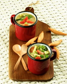 Delicious Homemade Vegetables Creamy Soup with Broccoli, Carrots, Zucchini, Leek, Red Bell Pepper and Green Pea in Red Soup Cups with Wooden Spoon and Bread Sticks closeup Wicker background