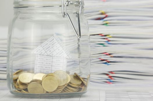 Close up blur house with stack of gold coins in bottle on finance account have calculator and overload of paperwork as background.