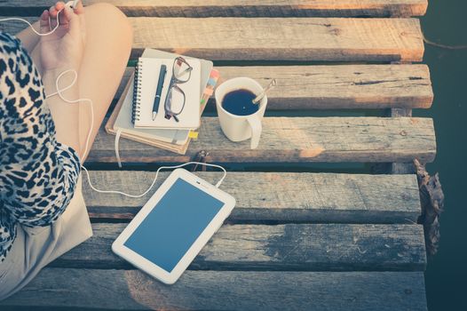 Young woman sitting on wood bridge with small tablet pc, coffee cup, glasses, and notebooks while listening to music in morning time with vintage filter effect