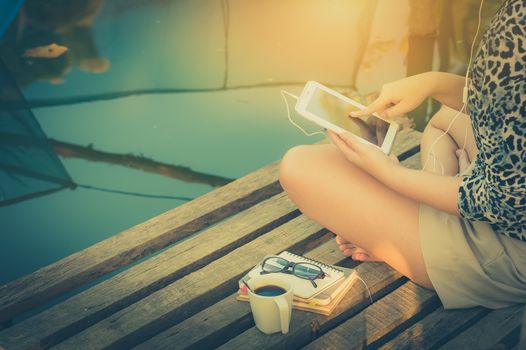 Young woman sitting on wood bridge touching on small talet pc screen while listening to music in morning time on weekend with vintage filter effect