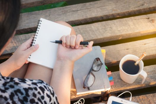 Young hipster woman writing journal on small notebook while sitting on wood bridge in morning time on weekend