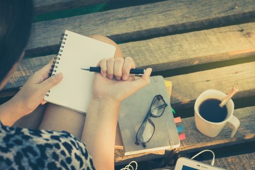 Young hipster woman writing journal on small notebook while sitting on wood bridge in morning time on weekend with vintage filter effect