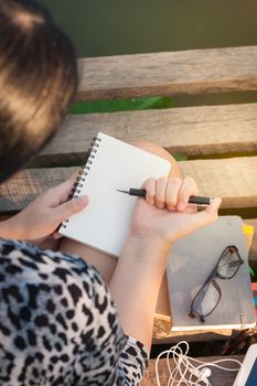 Young hipster woman writing journal on small notebook while sitting on wood bridge in morning time on weekend