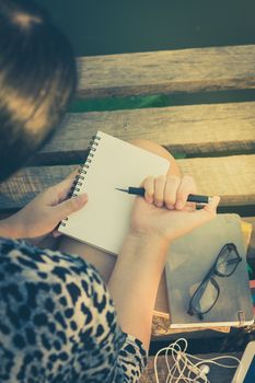Young hipster woman writing journal on small notebook while sitting on wood bridge in morning time on weekend with vintage filter effect