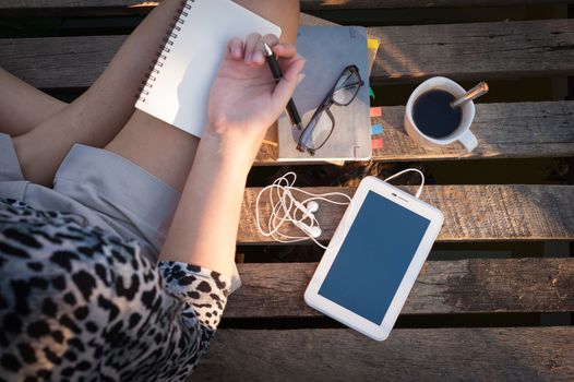 Small tablet pc is put down on wood bridge beside young woman, coffee cup, glasses, and notebooks in early morning time, focus on small tablet pc and notebooks