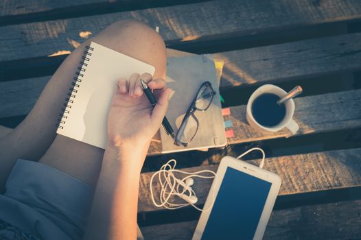 Young hipster woman writing journal on small notebook while sitting on wood litter in early morning time on weekend with vintage filter effect