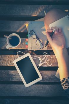 Small tablet pc is put down on wood bridge beside young woman, coffee cup, glasses, and notebooks on morning time with vintage filter effect