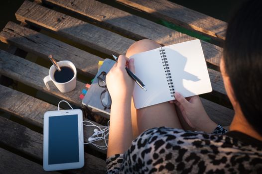 Young hipster woman writing journal on small notebook while sitting on wood bridge in early morning time on weekend with high contrast sun lighting