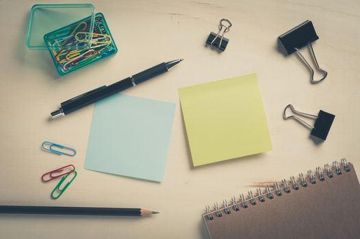 Sticky paper including blue, and green color with blank area for text or message stick on wood table with pen, pencil, paper clips, and notebook in morning time with vintage filter effect