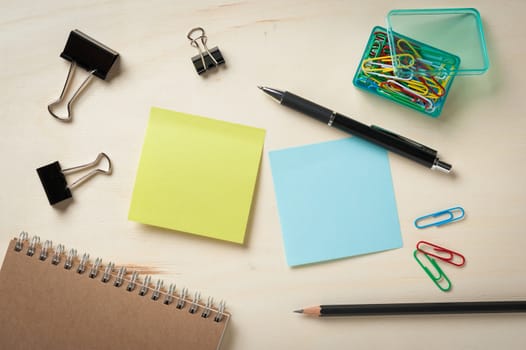 Sticky paper including blue, and green color with blank area for text or message stick on wood table with pen, pencil, paper clips, and notebook in morning time