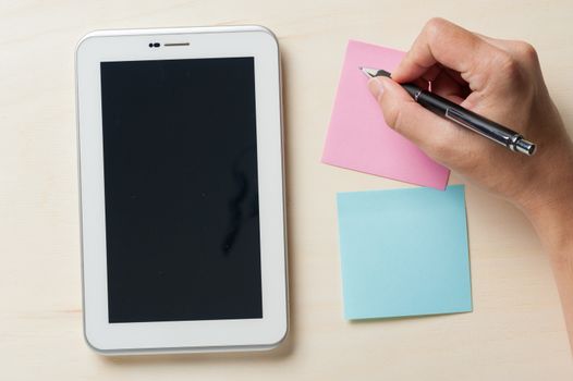 Young man writing with right hand on pink sticky paper beside small tablet pc with blank area on touchscreen on wood table