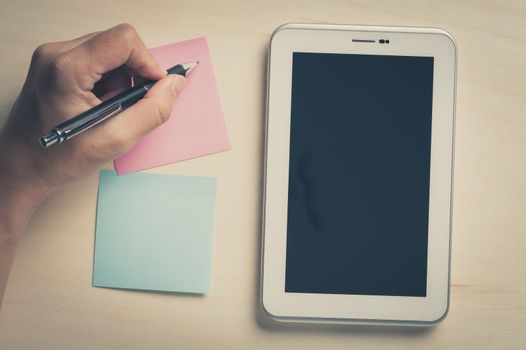 Young man writing with left hand on pink sticky paper beside small tablet pc with blank area on touchscreen on wood table with vintage filter effect