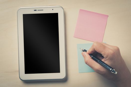 Young man writing with right hand on blue sticky paper beside small tablet pc with blank area on touchscreen on wood table with vintage filter effect