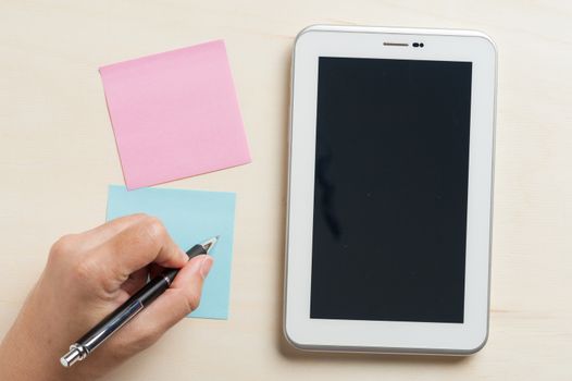 Young man writing with left hand on blue sticky paper beside small tablet pc with blank area on touchscreen on wood table