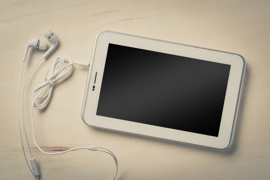 Small tablet pc with blank area on touchscreen and in ear headset on wood table with vintage filter effect